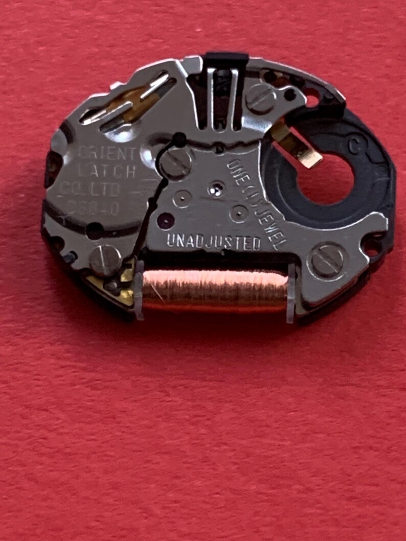 May include: Close-up of a watch movement with intricate metal components. The mechanism features the text "ORIENT LATCH CO. LTD" and "UNADJUSTED". A copper-colored coil is visible, along with a black plastic ring and a single jewel.