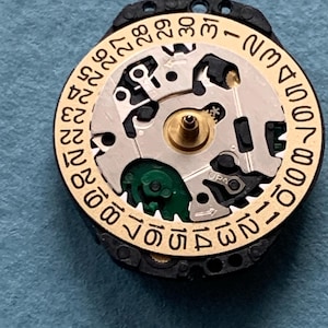 K&ouml;nnte beinhalten: Nahaufnahme eines Uhrwerks mit einem goldfarbenen Datumsrad und einem schwarzen Au&szlig;enring. Das Datumsrad hat Zahlen von 1 bis 31. Der Uhrenmechanismus ist silberfarben und hat kleine Zahnr&auml;der und Komponenten. Der Hintergrund ist blau.