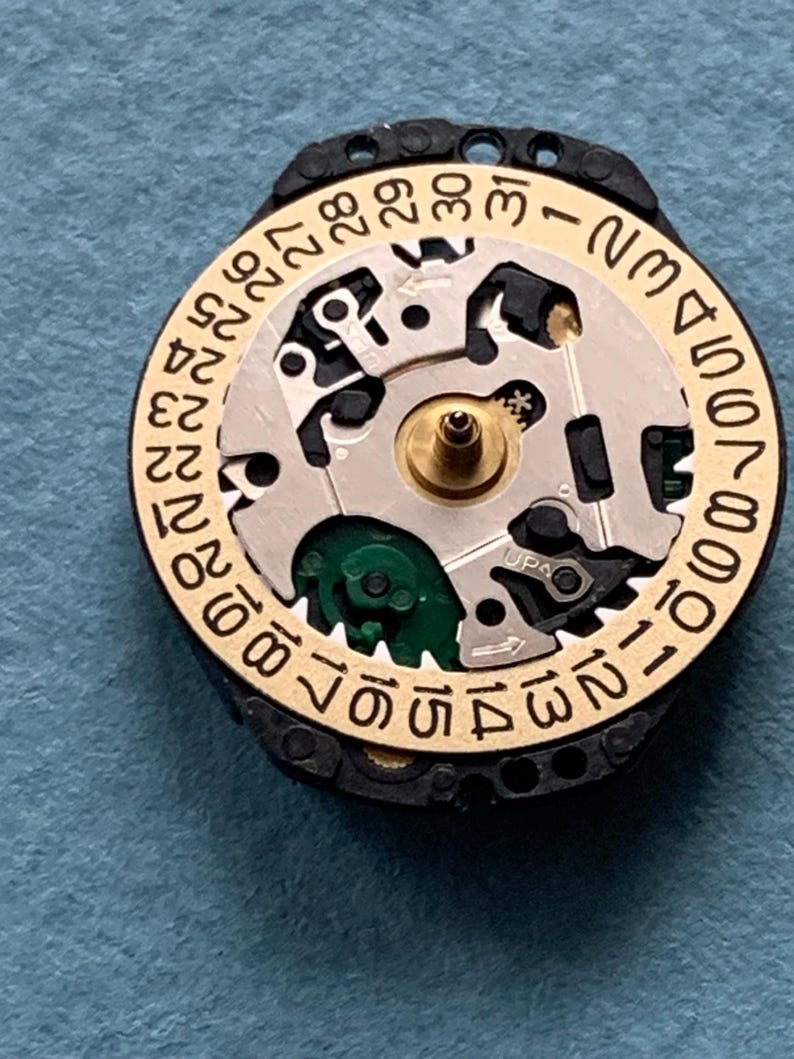 K&ouml;nnte beinhalten: Nahaufnahme eines Uhrwerks mit einem goldfarbenen Datumsrad und einem schwarzen Au&szlig;enring. Das Datumsrad hat Zahlen von 1 bis 31. Der Uhrenmechanismus ist silberfarben und hat kleine Zahnr&auml;der und Komponenten. Der Hintergrund ist blau.