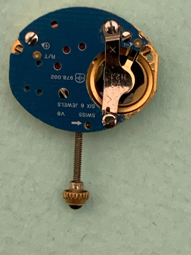 May include: A watch movement with a blue circuit board, gold-colored components, and a silver-colored screw. The board has text including "SWISS", "6 JEWELS", and "978.002". A gold-colored crown is at the bottom.