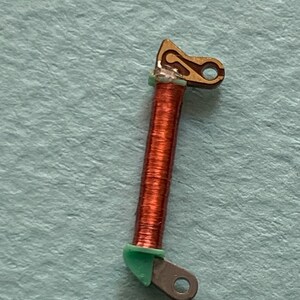 May include: Close-up of a small, copper-colored coil with a brown, gold, and green component. The coil is wrapped around a central core. The gold component has a curved design with a hole. The green component is at the base of the coil.