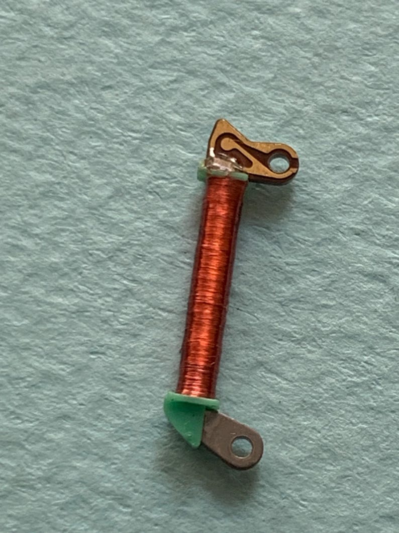 May include: Close-up of a small, copper-colored coil with a brown, gold, and green component. The coil is wrapped around a central core. The gold component has a curved design with a hole. The green component is at the base of the coil.