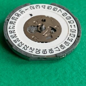May include: A watch movement component with a white date wheel featuring black numbers from 1 to 31. The component has a silver-colored metal frame and a central mechanism with small gears and screws. The background is a solid green.