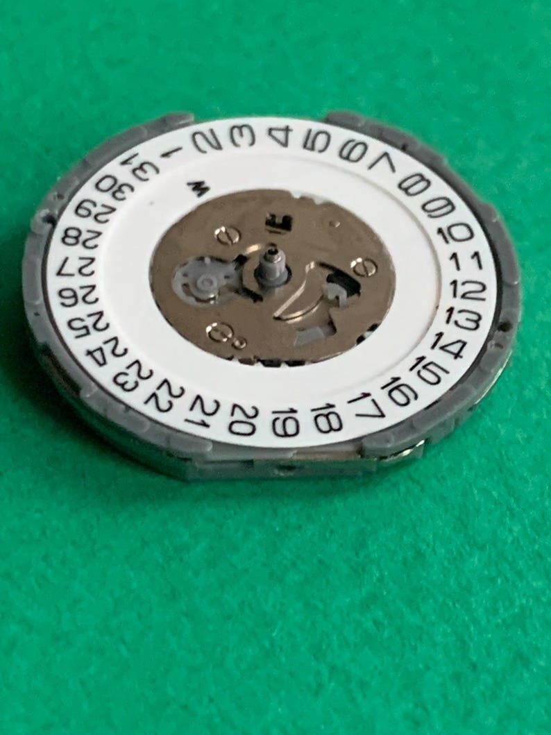 May include: A watch movement component with a white date wheel featuring black numbers from 1 to 31. The component has a silver-colored metal frame and a central mechanism with small gears and screws. The background is a solid green.