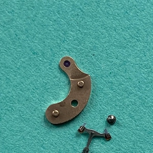 May include: Close-up of watch parts on a teal surface. The parts include a curved, metallic piece with three holes and a purple dot, a small ball, and a tiny, intricate metal component with red accents.