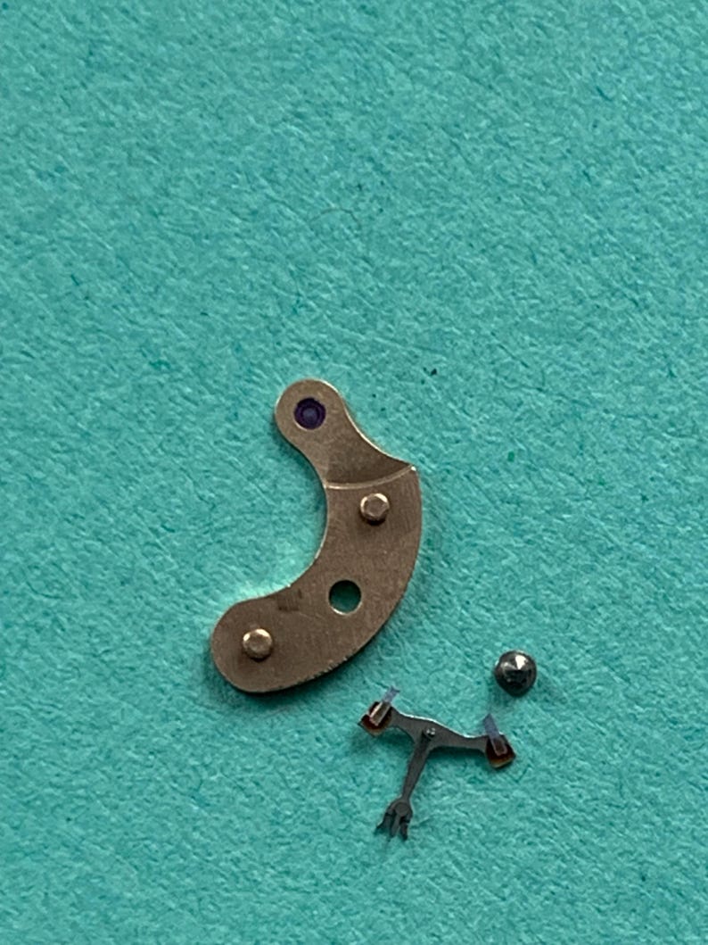 May include: Close-up of watch parts on a teal surface. The parts include a curved, metallic piece with three holes and a purple dot, a small ball, and a tiny, intricate metal component with red accents.