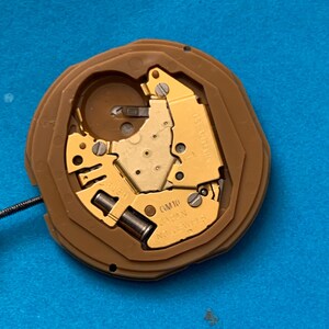 May include: A watch movement with a brown plastic casing and gold-colored internal components. The movement has the text "GM10 JAPAN" and "NO JEWELS" printed on it. A thin wire extends from the side. The background is a solid blue color.