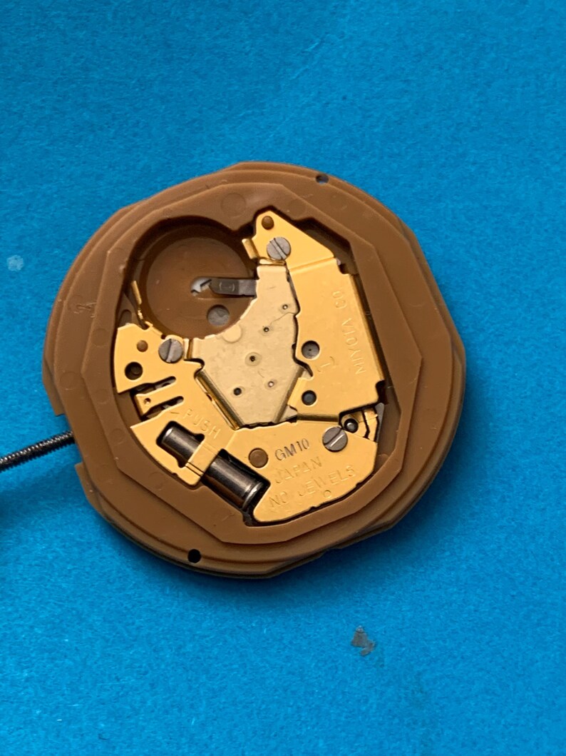 May include: A watch movement with a brown plastic casing and gold-colored internal components. The movement has the text "GM10 JAPAN" and "NO JEWELS" printed on it. A thin wire extends from the side. The background is a solid blue color.