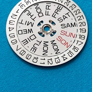 May include: A circular watch calendar dial with the days of the week and dates printed on it. The days are in English, with "SUN" in red. The dial has a central hole and is set against a blue background.