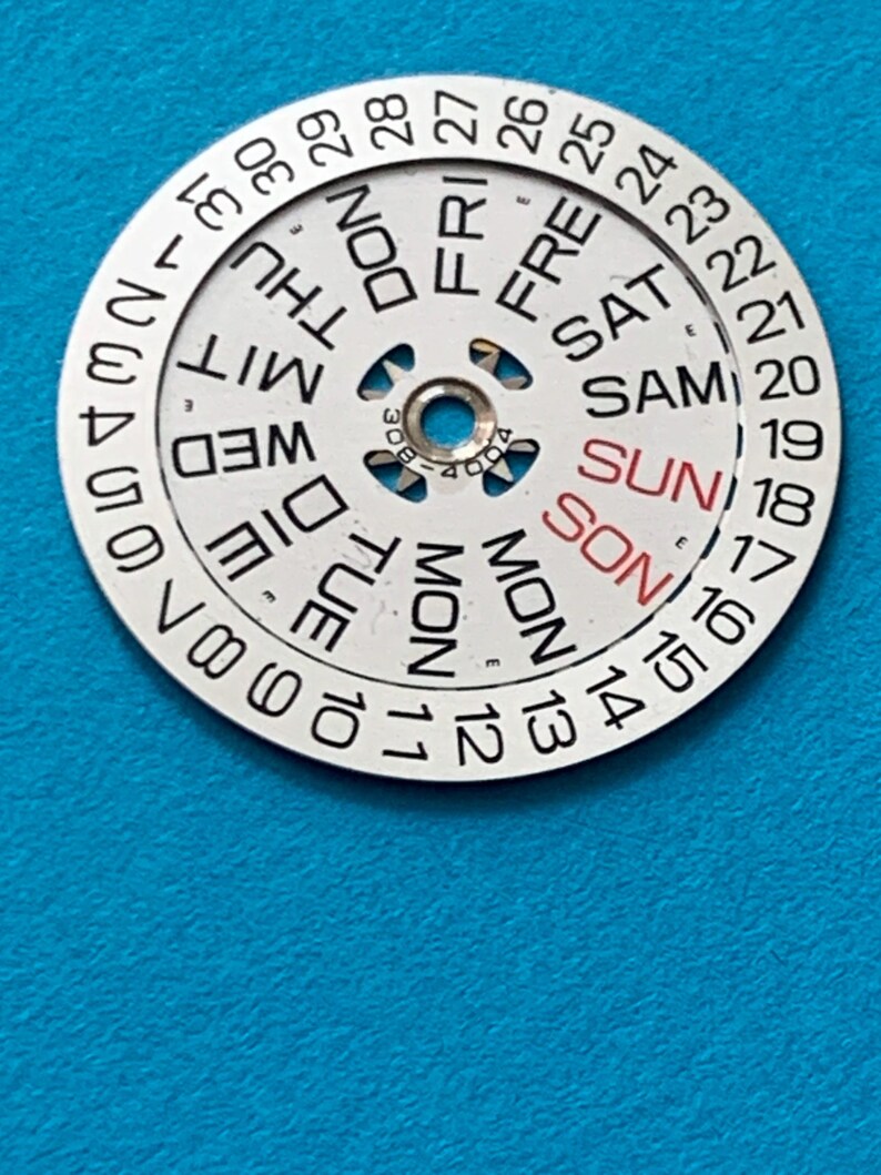 May include: A circular watch calendar dial with the days of the week and dates printed on it. The days are in English, with "SUN" in red. The dial has a central hole and is set against a blue background.