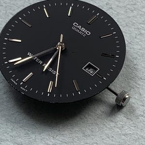 Puede incluir: Una esfera de reloj Casio Quartz negra con manecillas y marcadores dorados. La esfera del reloj tiene una ventana de fecha y una corona plateada. El texto "CASIO QUARTZ" y "WATER RESIST" son visibles.