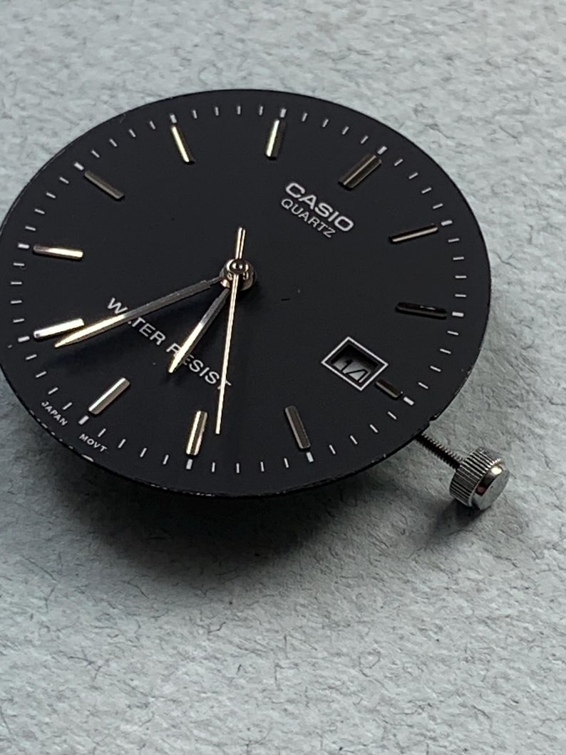Puede incluir: Una esfera de reloj Casio Quartz negra con manecillas y marcadores dorados. La esfera del reloj tiene una ventana de fecha y una corona plateada. El texto "CASIO QUARTZ" y "WATER RESIST" son visibles.