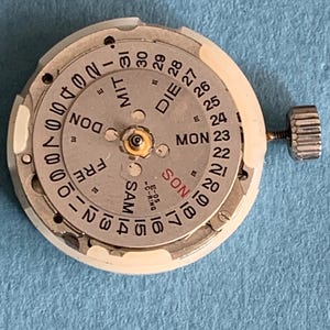 May include: A watch movement with a silver dial and a white outer ring. The dial displays the days of the week and the date, with a small crown on the side. The text includes "MON", "DIE", "FRE", "SAM", and numbers.