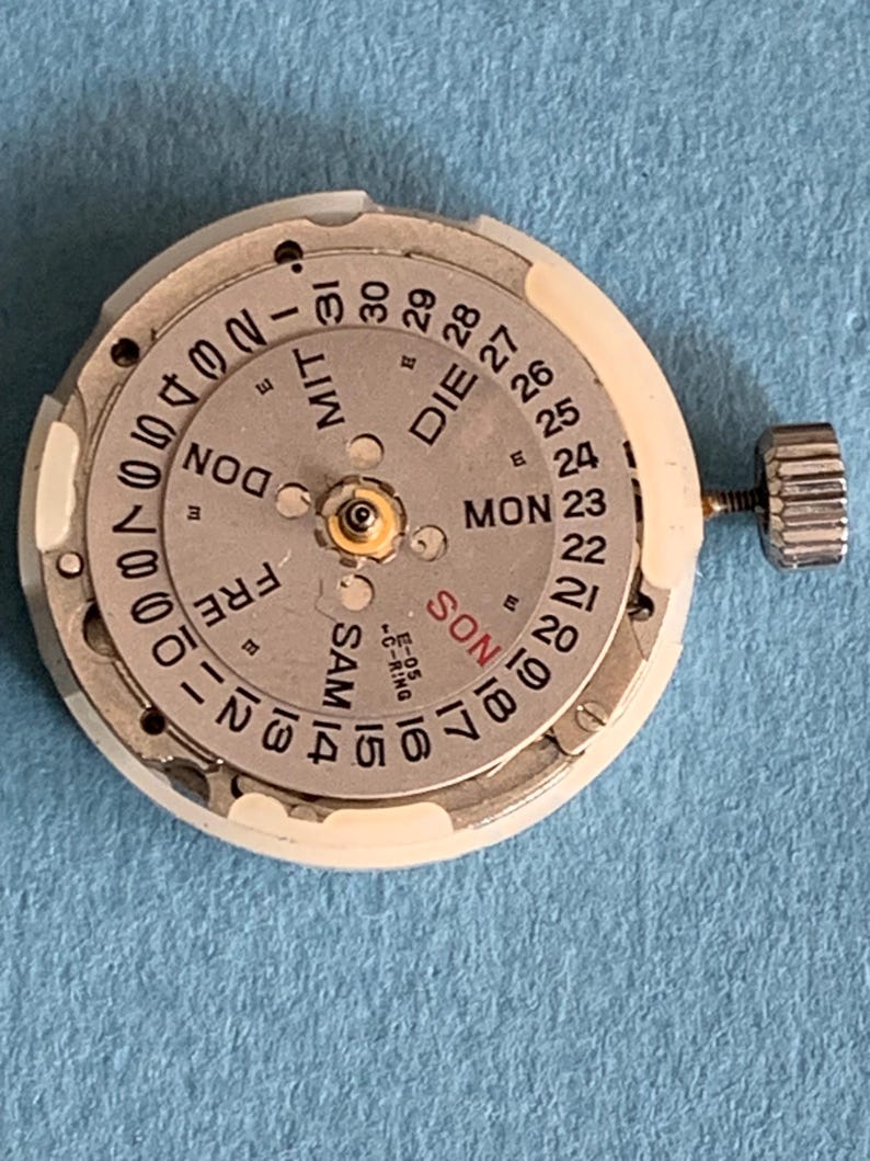 May include: A watch movement with a silver dial and a white outer ring. The dial displays the days of the week and the date, with a small crown on the side. The text includes "MON", "DIE", "FRE", "SAM", and numbers.