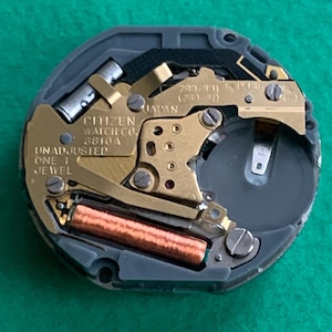 May include: A close-up of a Citizen watch movement, featuring gold-toned gears and components set within a gray casing. The text "CITIZEN WATCH CO. 3810A JAPAN" is visible. The watch mechanism includes a battery and a copper-colored coil.