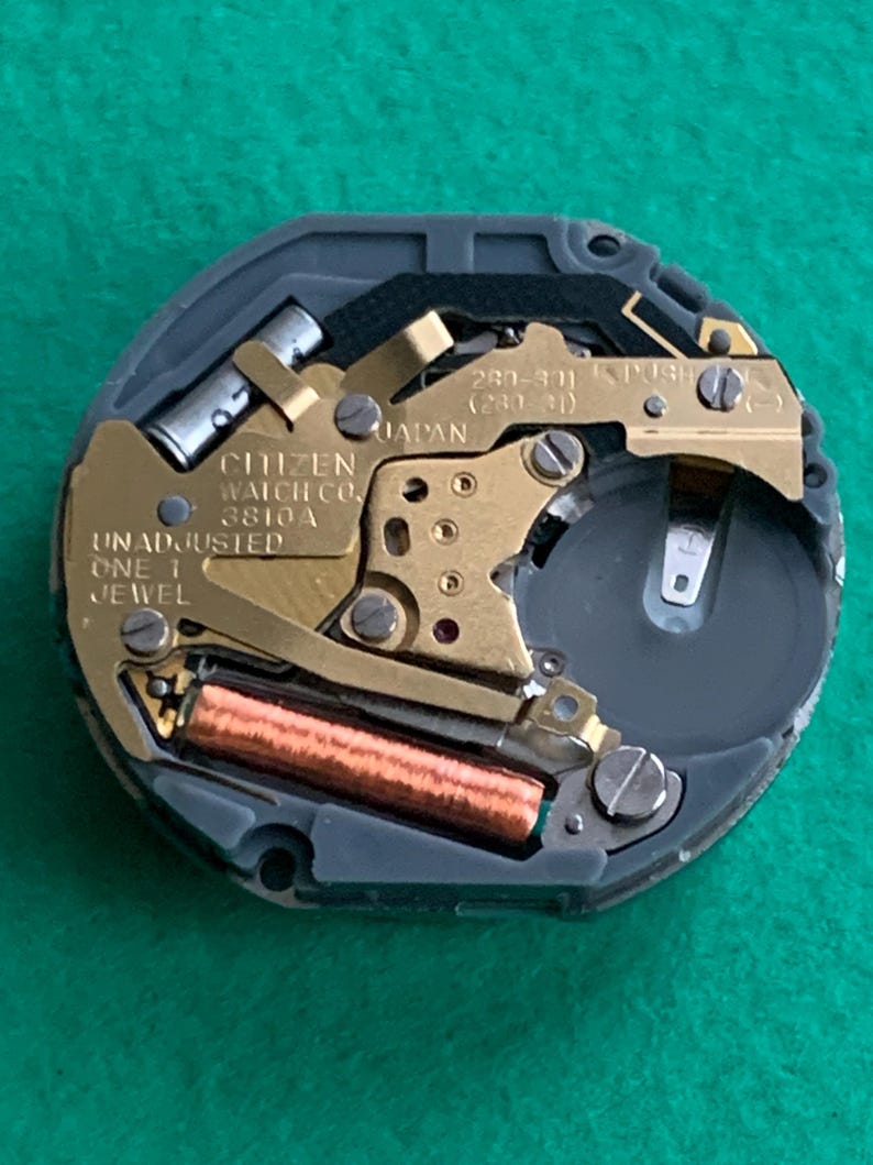 May include: A close-up of a Citizen watch movement, featuring gold-toned gears and components set within a gray casing. The text "CITIZEN WATCH CO. 3810A JAPAN" is visible. The watch mechanism includes a battery and a copper-colored coil.