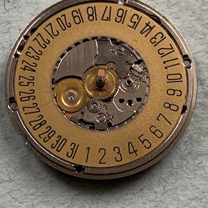May include: Close-up of a watch movement with a gold-colored dial and silver-colored metal casing. The dial features the numbers 1 through 31, indicating the days of the month. The intricate gears and mechanisms of the watch are visible.
