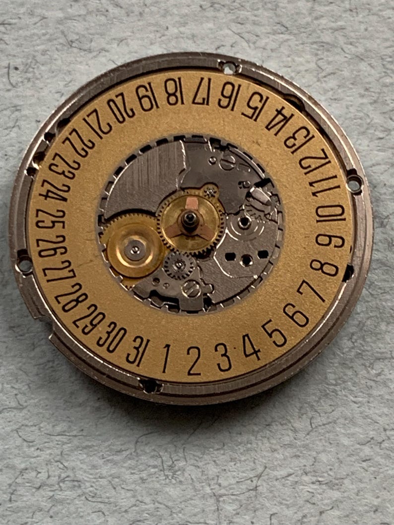 May include: Close-up of a watch movement with a gold-colored dial and silver-colored metal casing. The dial features the numbers 1 through 31, indicating the days of the month. The intricate gears and mechanisms of the watch are visible.