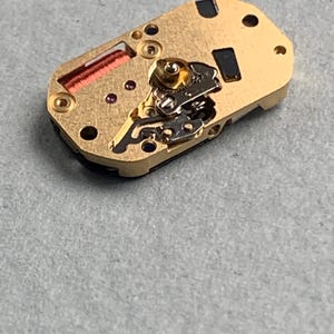 May include: A gold-toned watch movement with intricate gears, screws, and a red coil. The rectangular component has several small holes and black rectangular insets. The watch mechanism is set against a gray surface.