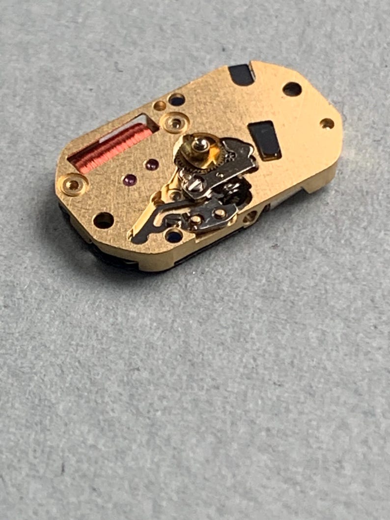 May include: A gold-toned watch movement with intricate gears, screws, and a red coil. The rectangular component has several small holes and black rectangular insets. The watch mechanism is set against a gray surface.