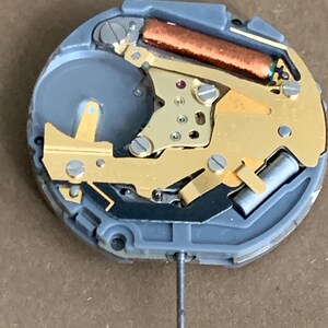 May include: Close-up of a watch movement with a gray plastic base and gold-colored metal components. A copper-colored cylinder and a silver metal pin are also visible. The watch mechanism is designed for timekeeping.