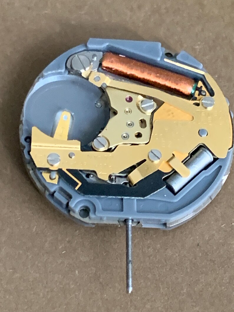May include: Close-up of a watch movement with a gray plastic base and gold-colored metal components. A copper-colored cylinder and a silver metal pin are also visible. The watch mechanism is designed for timekeeping.
