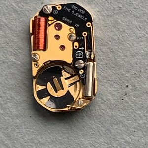 May include: Close-up of a watch movement with gold-colored components, including a coil, gears, and screws. The black plate has the text "280 002 FIVE 5 JEWELS SWISS V8". The watch mechanism is on a gray surface.