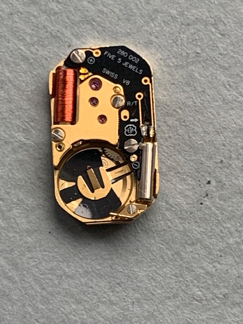 May include: Close-up of a watch movement with gold-colored components, including a coil, gears, and screws. The black plate has the text "280 002 FIVE 5 JEWELS SWISS V8". The watch mechanism is on a gray surface.