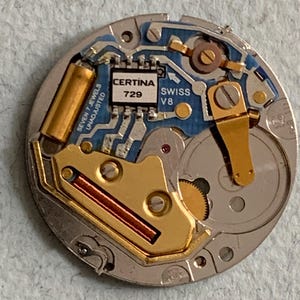 Może przedstawiać: Zbliżenie mechanizmu zegarka Certina. Mechanizm zawiera złote elementy, niebieską płytkę obwodu drukowanego oraz napis "CERTINA 729 SWISS V8". Okrągły mechanizm ma małe śruby i koła zębate.