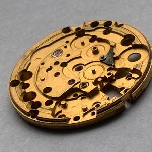 May include: Close-up of a gold-colored watch movement. The intricate mechanism features various gears, holes, and a small ruby-colored jewel. The watch movement is oval-shaped and appears to be vintage.