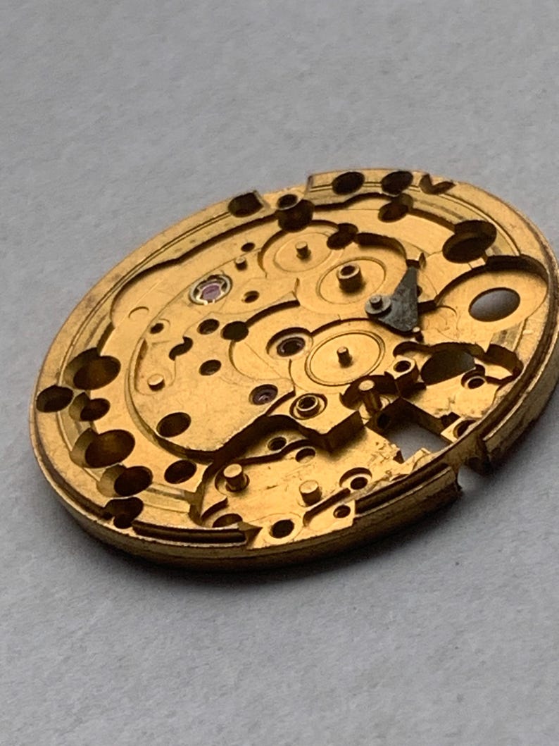 May include: Close-up of a gold-colored watch movement. The intricate mechanism features various gears, holes, and a small ruby-colored jewel. The watch movement is oval-shaped and appears to be vintage.