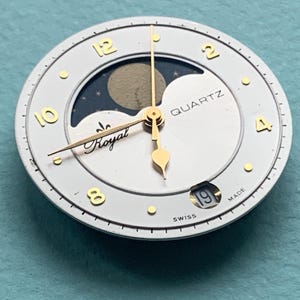 Op de afbeelding: Een witte wijzerplaat met goudkleurige wijzers en cijfers. De wijzerplaat heeft een maanfase-indicator en het woord "QUARTZ". De tekst "Royal" is ook zichtbaar. De datum wordt onderaan weergegeven. De wijzerplaat is gemarkeerd met "SWISS MADE".