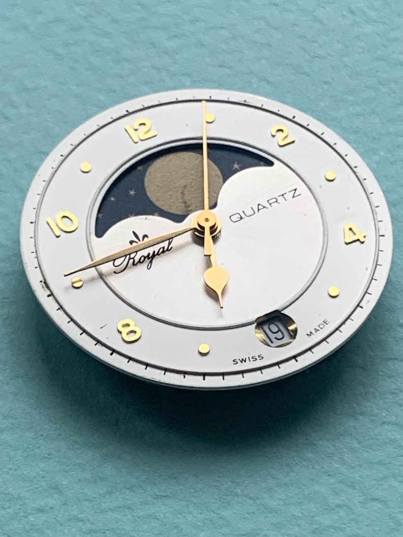 Op de afbeelding: Een witte wijzerplaat met goudkleurige wijzers en cijfers. De wijzerplaat heeft een maanfase-indicator en het woord "QUARTZ". De tekst "Royal" is ook zichtbaar. De datum wordt onderaan weergegeven. De wijzerplaat is gemarkeerd met "SWISS MADE".