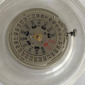 May include: Close-up of a watch movement with a silver-colored dial and a calendar function. The dial displays the days of the week and the date, with a small crown on the side. The movement is encased in a clear plastic container.