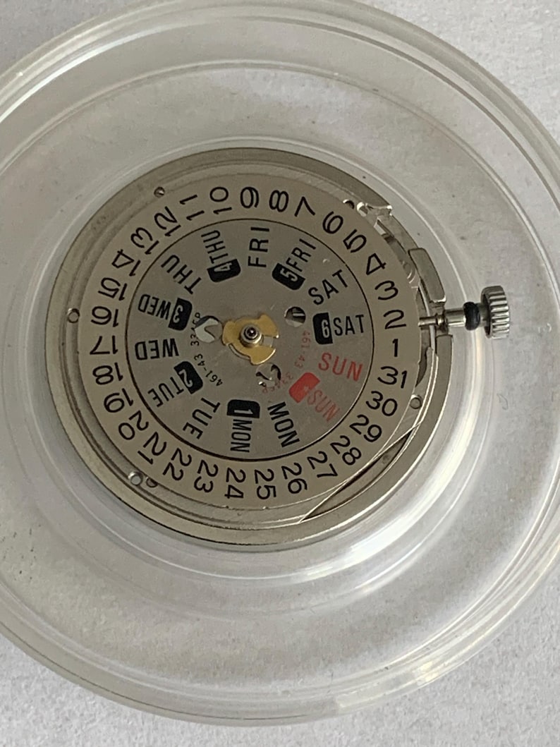 May include: Close-up of a watch movement with a silver-colored dial and a calendar function. The dial displays the days of the week and the date, with a small crown on the side. The movement is encased in a clear plastic container.