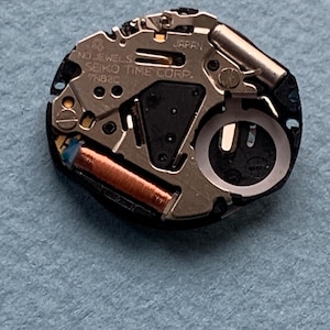 K&ouml;nnte beinhalten: Nahaufnahme eines Uhrwerks mit dem Text "JAPAN" und "SEIKO TIME CORP." sichtbar. Das Uhrwerk weist verschiedene metallische Komponenten auf, darunter eine Kupferspule und eine silberfarbene Unruh. Der Hintergrund ist ein hellblauer Stoff.