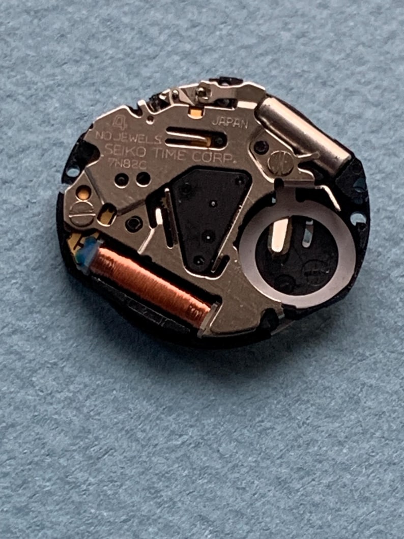 K&ouml;nnte beinhalten: Nahaufnahme eines Uhrwerks mit dem Text "JAPAN" und "SEIKO TIME CORP." sichtbar. Das Uhrwerk weist verschiedene metallische Komponenten auf, darunter eine Kupferspule und eine silberfarbene Unruh. Der Hintergrund ist ein hellblauer Stoff.