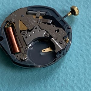 K&ouml;nnte beinhalten: Nahaufnahme eines Uhrwerks mit komplexen Zahnr&auml;dern, einer Kupferspule und einer goldfarbenen Krone. Der Mechanismus ist grau und schwarz, mit goldenen und silberfarbenen Komponenten. Der Text "ONE JEWEL" ist sichtbar.