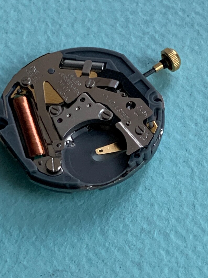 K&ouml;nnte beinhalten: Nahaufnahme eines Uhrwerks mit komplexen Zahnr&auml;dern, einer Kupferspule und einer goldfarbenen Krone. Der Mechanismus ist grau und schwarz, mit goldenen und silberfarbenen Komponenten. Der Text "ONE JEWEL" ist sichtbar.