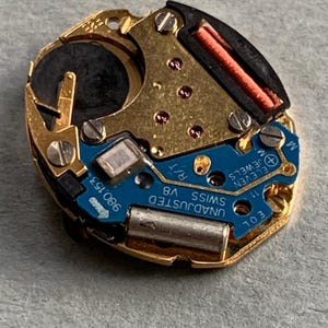 May include: A detailed view of a watch movement featuring gold-toned metal parts, a blue circuit board, and tiny screws. The movement is marked with "UNADJUSTED SWISS V8" and "ELEVEN JEWELS". A black battery and a red coil are also present.