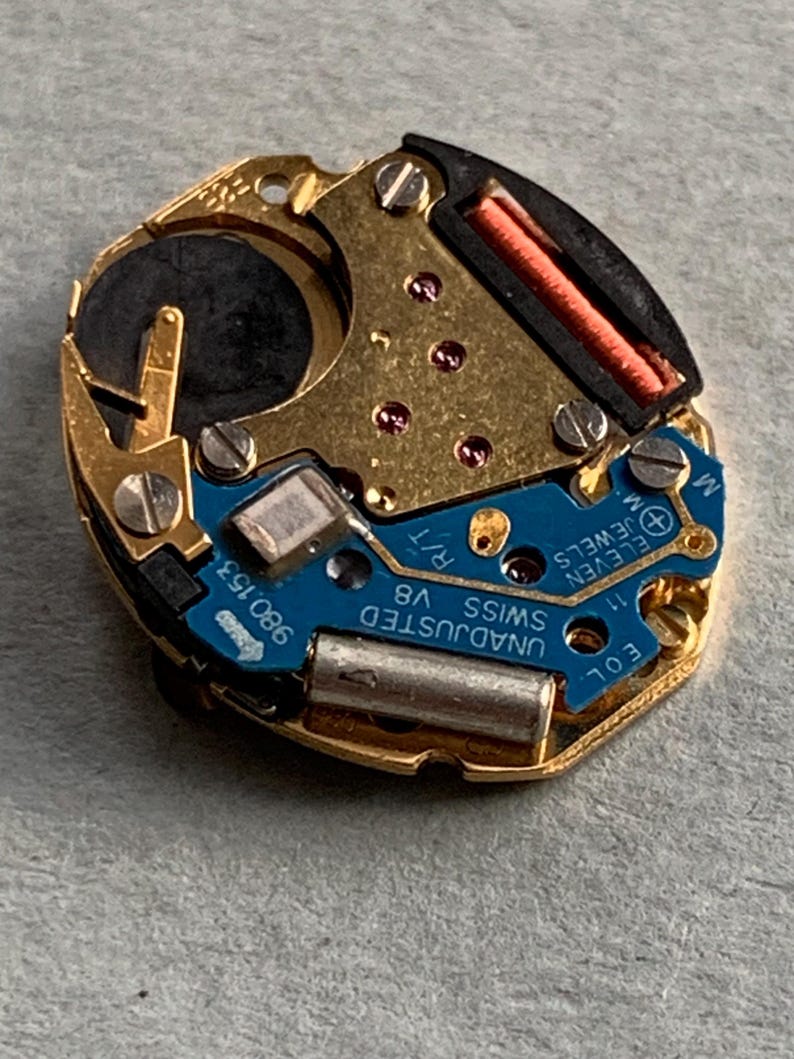 May include: A detailed view of a watch movement featuring gold-toned metal parts, a blue circuit board, and tiny screws. The movement is marked with "UNADJUSTED SWISS V8" and "ELEVEN JEWELS". A black battery and a red coil are also present.