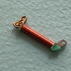 May include: A small, detailed electronic component featuring a copper coil wrapped around a central core. The component has gold-colored and silver-colored metal ends, with green insulation at the connection points. The background is a light blue surface.