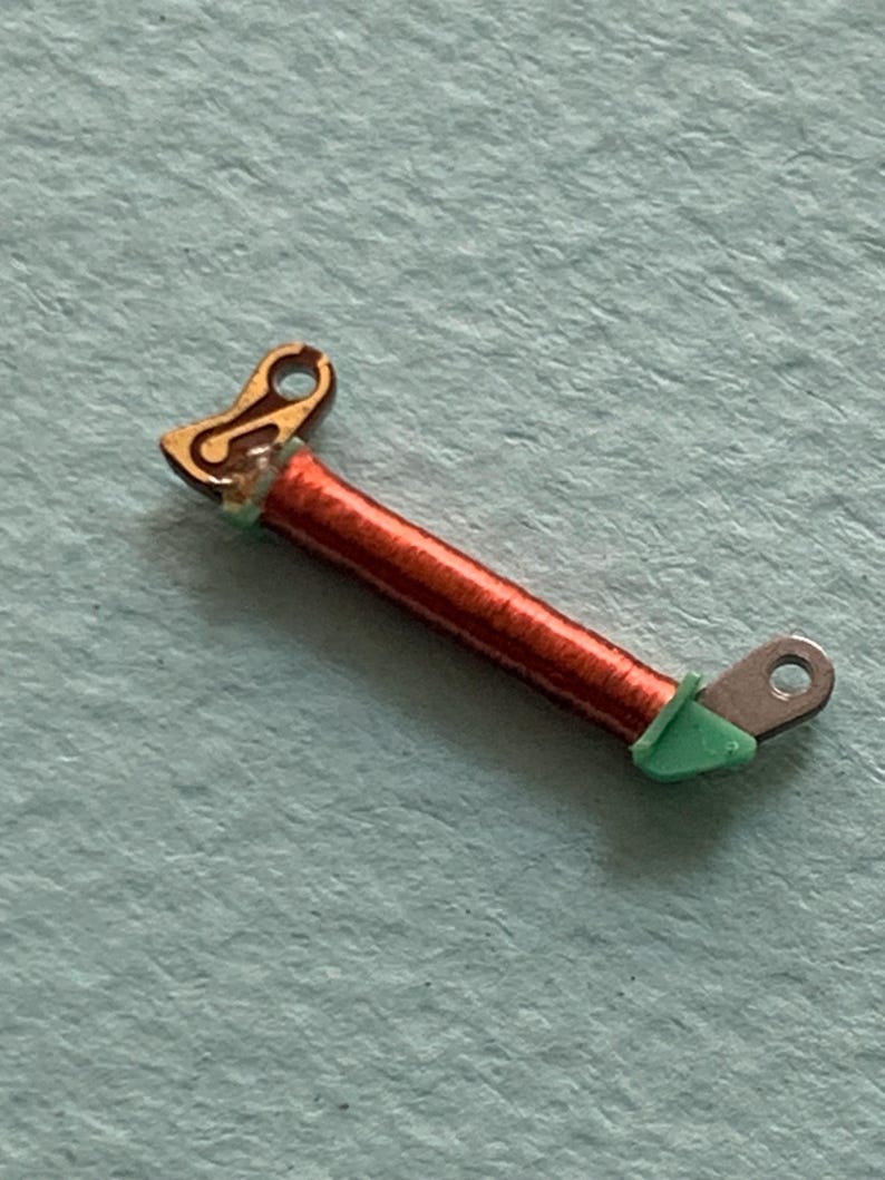 May include: A small, detailed electronic component featuring a copper coil wrapped around a central core. The component has gold-colored and silver-colored metal ends, with green insulation at the connection points. The background is a light blue surface.