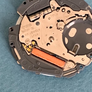 May include: Close-up of a watch movement with intricate gears and components. The watch mechanism features a silver-colored metal plate with the text "CITIZEN WATCH CO. NO JEWELS JAPAN". An orange rectangular component is visible.