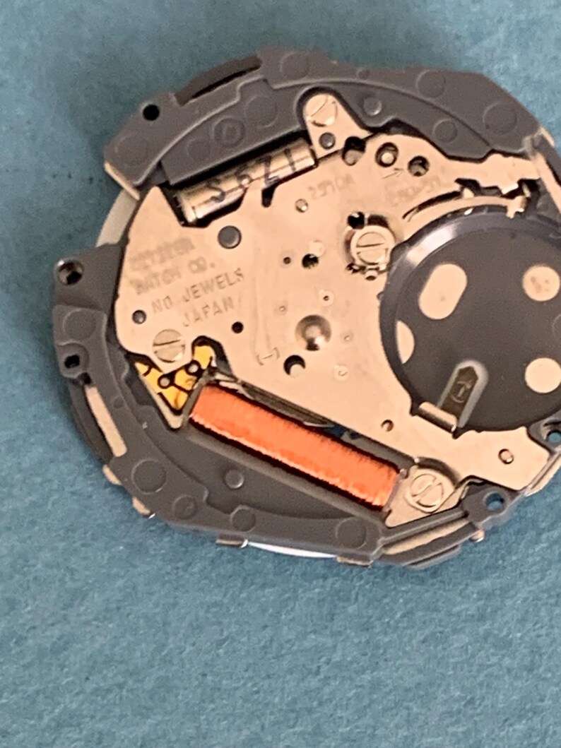 May include: Close-up of a watch movement with intricate gears and components. The watch mechanism features a silver-colored metal plate with the text "CITIZEN WATCH CO. NO JEWELS JAPAN". An orange rectangular component is visible.