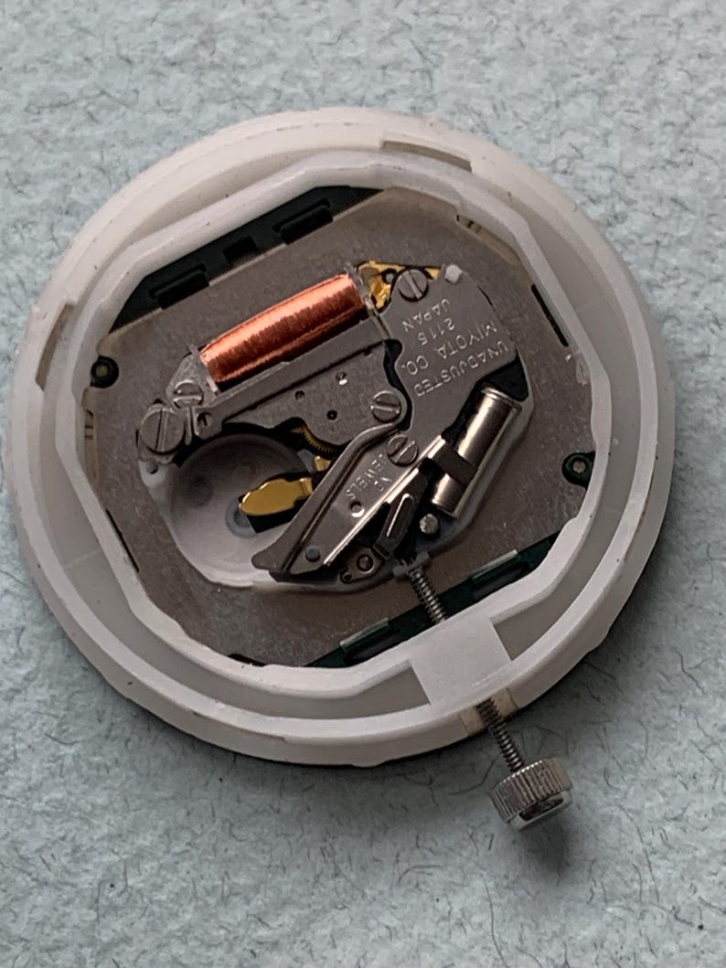 Puede incluir: Primer plano de un movimiento de reloj, con un mecanismo plateado y cobrizo dentro de una carcasa de pl&aacute;stico blanco. El movimiento incluye el texto "MIYOTA CO. JAPAN 2115 UNADJUSTED". Se ve un tornillo plateado en el lateral.