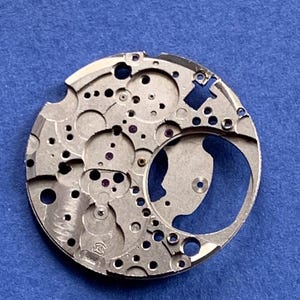 Puede incluir: Primer plano de un movimiento de reloj plateado con intrincados engranajes, agujeros y peque&ntilde;as joyas moradas. El mecanismo circular se encuentra sobre un fondo azul, mostrando su dise&ntilde;o detallado y artesan&iacute;a.