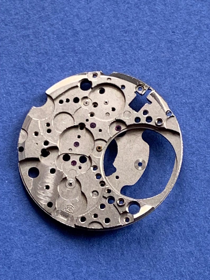 Puede incluir: Primer plano de un movimiento de reloj plateado con intrincados engranajes, agujeros y peque&ntilde;as joyas moradas. El mecanismo circular se encuentra sobre un fondo azul, mostrando su dise&ntilde;o detallado y artesan&iacute;a.
