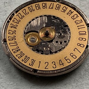 May include: Close-up of a watch movement with a gold-colored date wheel. The wheel has numbers from 1 to 31, indicating the days of the month. The intricate mechanical parts are visible.