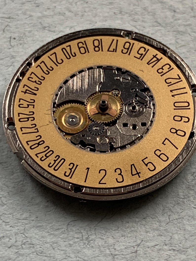 May include: Close-up of a watch movement with a gold-colored date wheel. The wheel has numbers from 1 to 31, indicating the days of the month. The intricate mechanical parts are visible.